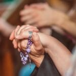 Kiedy różaniec staje się rutyną: sposoby na żywą modlitwę tajemnicami Złożone dłonie w modlitwie trzymające fioletowy różaniec w kościele